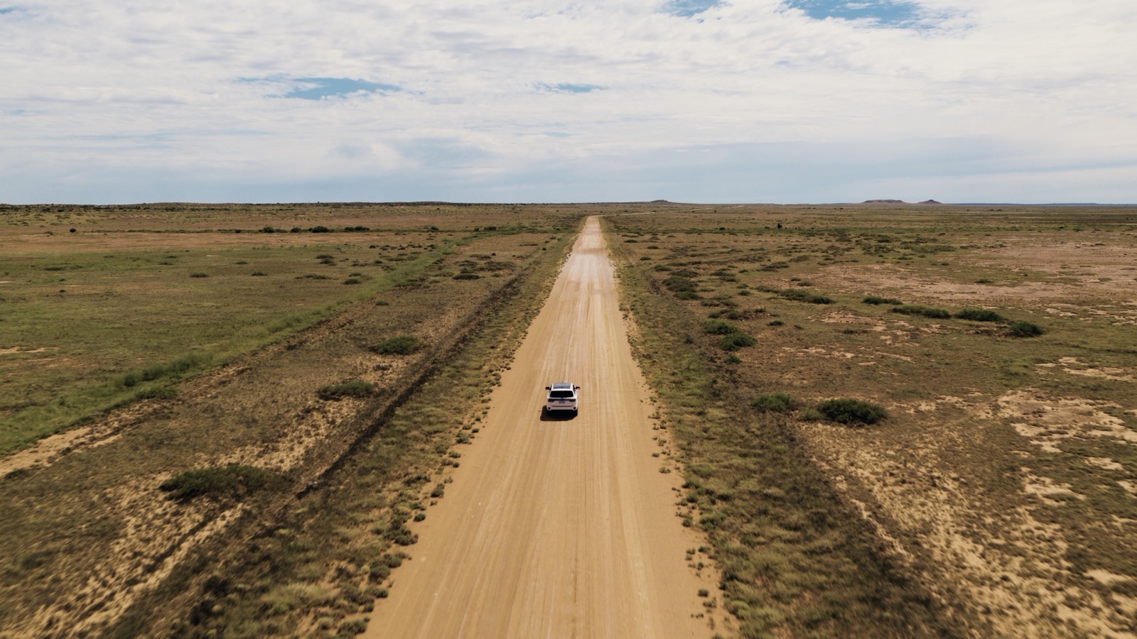 Graded: aerial of a white 4WD on a straight gravel track through low scrub